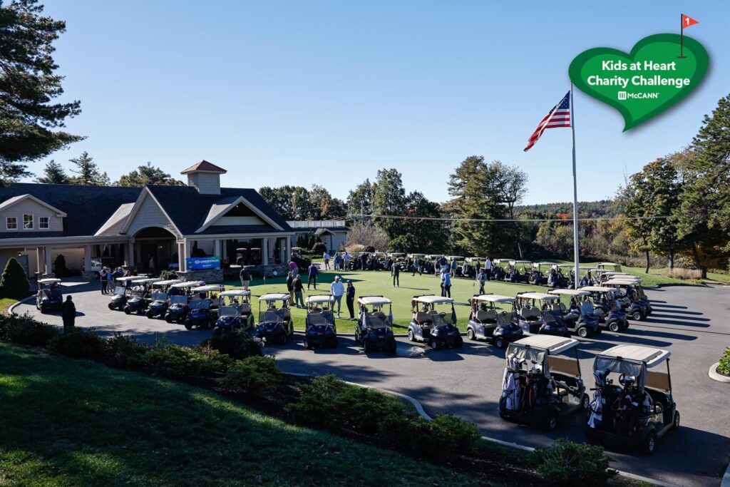 A view of a golf course with a large group of golf carts lined up outside a clubhouse, with people gathered near the entrance and an American flag flying on a tall flagpole under a clear sky.