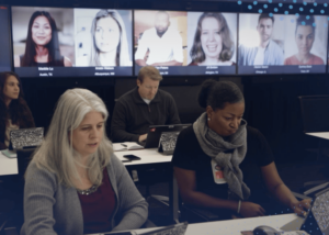 A classroom with four students using tablets and a large screen behind them displaying the icons of those on video call.