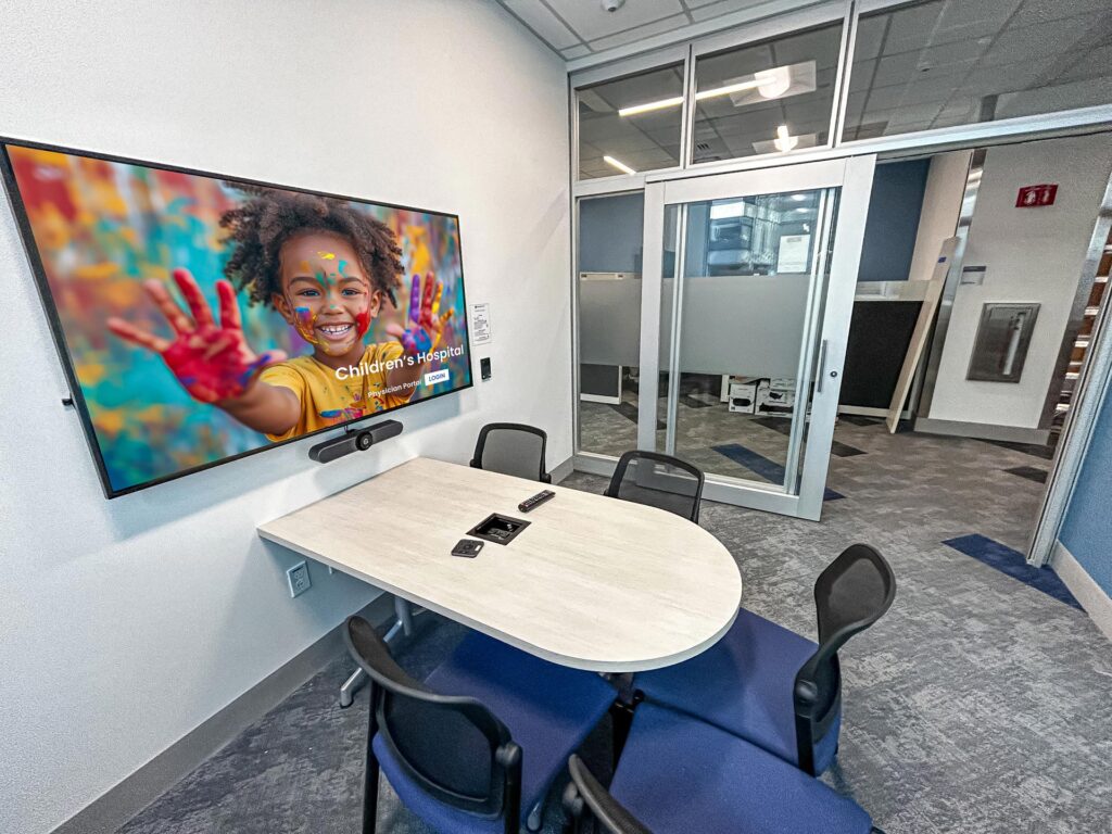 A small, modern conference room with a mounted screen displaying an image of a smiling child with painted hands. A video conferencing camera is installed below the screen, and a sliding glass door leads to an open office space.