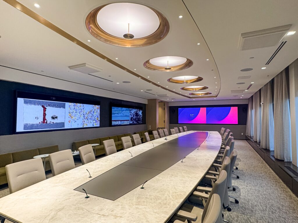 Executive Briefing Center designed for high-profile meetings, strategic planning, and leadership discussions. The space features a state-of-the-art boardroom with an elegant marble conference table, integrated microphones, and advanced AV capabilities. Wall-mounted displays showcase dynamic content, including artwork and corporate presentations, creating an immersive environment for executive decision-making. The sophisticated ceiling design, ambient lighting, and floor-to-ceiling curtains enhance the luxurious and modern atmosphere of the space, reflecting McCann Systems' expertise in AV integration and future-proof communication solutions.