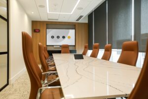 A modern conference room with a long marble table, brown faux leather chairs, and a video display on the back wall.