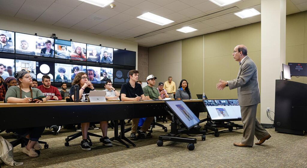 Professor speaking to students in suit, with video wall behind showing remote participants
