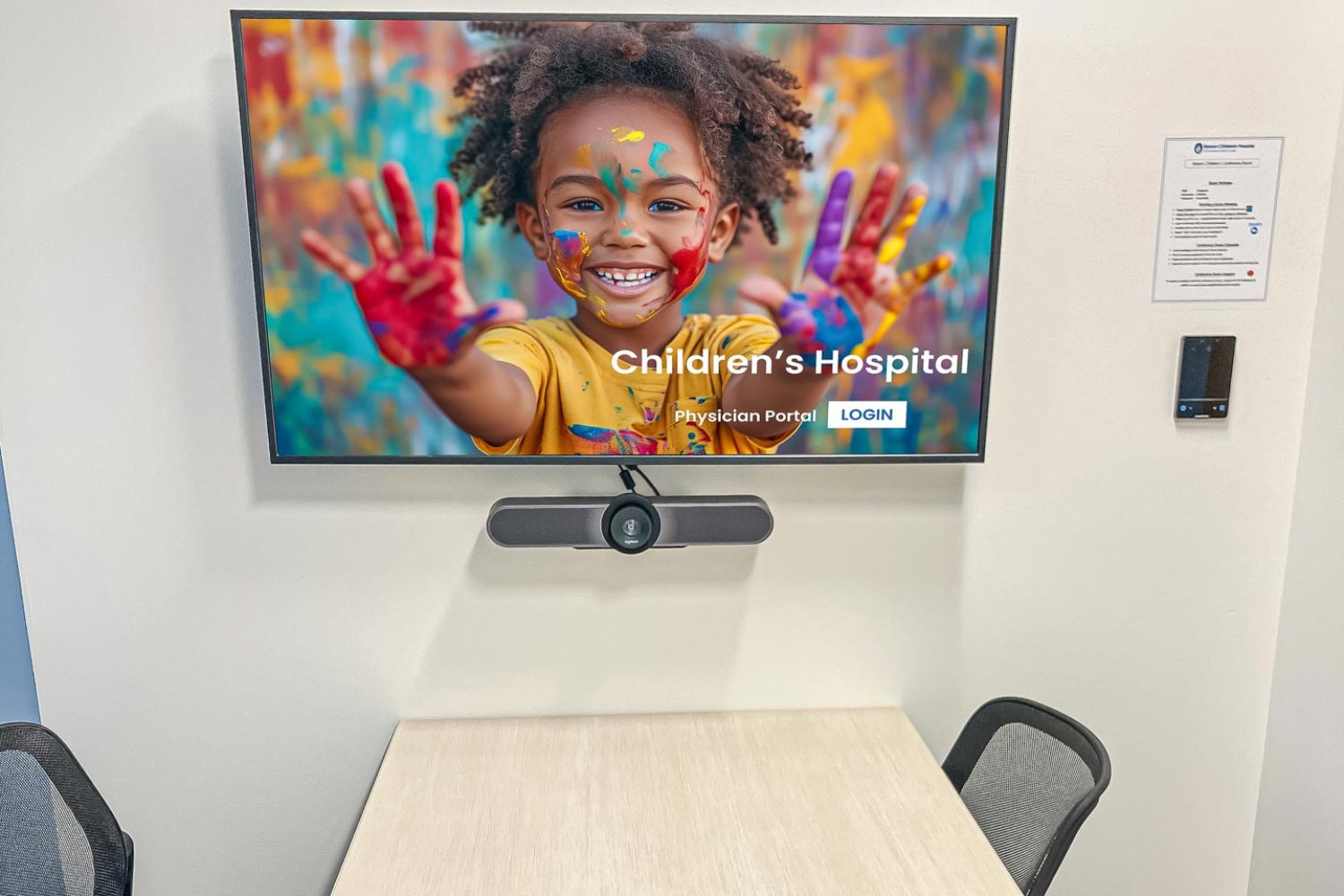 A small meeting room with a wooden conference table, four chairs, and a wall-mounted screen displaying an image of a smiling child with painted hands. A video conferencing camera is installed below the screen.