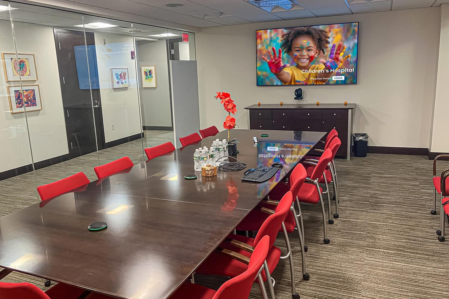 A spacious conference room with a large dark wooden table surrounded by red chairs. A wall-mounted screen displays a colorful image of a smiling child with painted hands. A glass wall separates the room from an adjacent hallway.