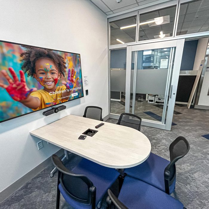 A small, modern conference room with a mounted screen displaying an image of a smiling child with painted hands. A video conferencing camera is installed below the screen, and a sliding glass door leads to an open office space.