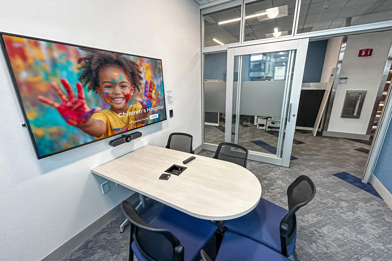 A small, modern conference room with a mounted screen displaying an image of a smiling child with painted hands. A video conferencing camera is installed below the screen, and a sliding glass door leads to an open office space.