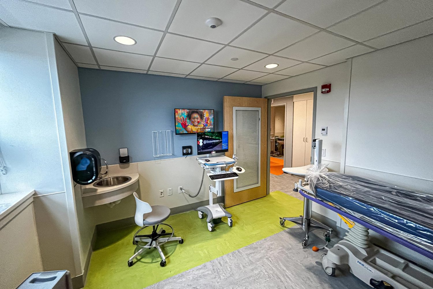 A medical examination room with a mounted screen on the wall displaying an image of a smiling child with painted hands. A rolling workstation, sink, and an examination table with protective plastic covering are present.