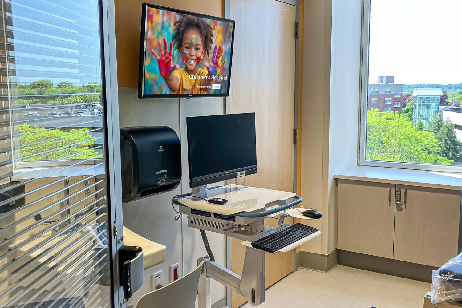 A hospital room with a mounted screen displaying a smiling child with painted hands. A rolling workstation and an adjustable chair are placed near a large window with a view of trees and buildings.