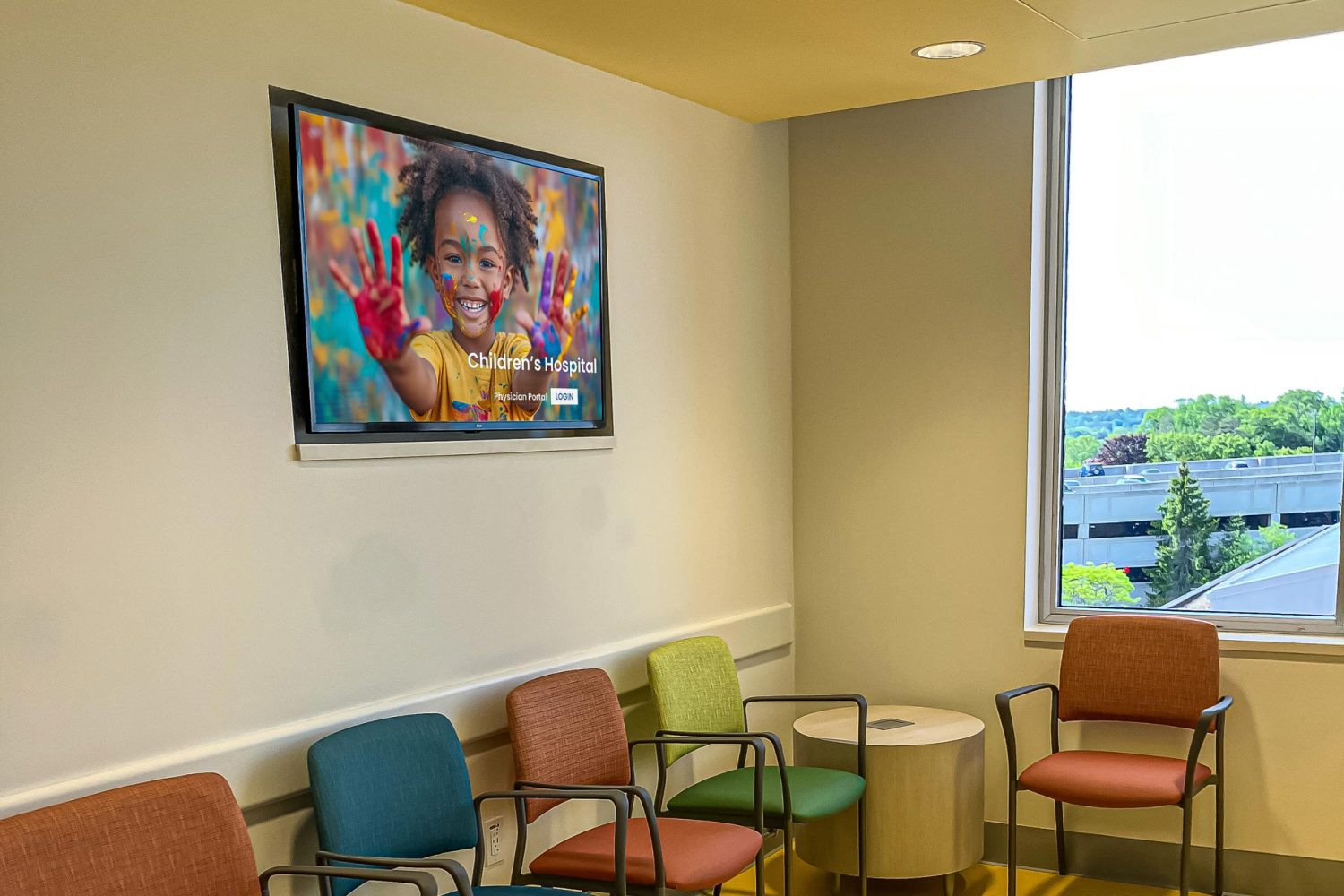 A waiting area with colorful chairs arranged around a small table. A wall-mounted screen displays an image of a smiling child with painted hands, and a large window lets in natural light.