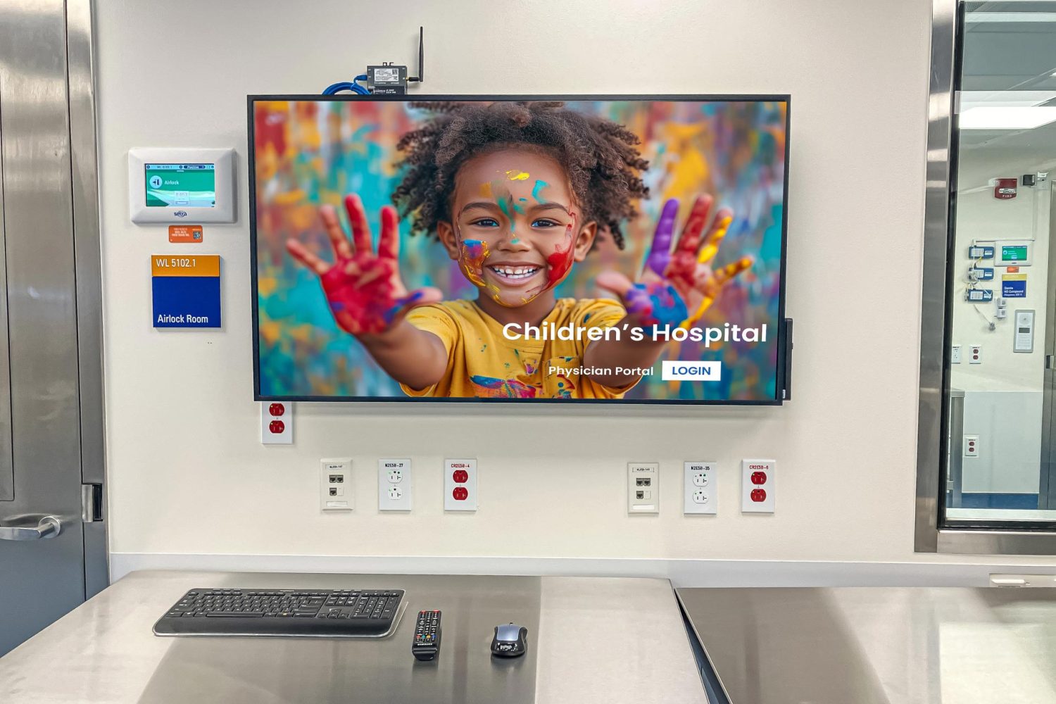 A sterile hospital room with a stainless steel workstation. A mounted screen on the wall displays an image of a smiling child with painted hands. Medical equipment and multiple power outlets are visible.