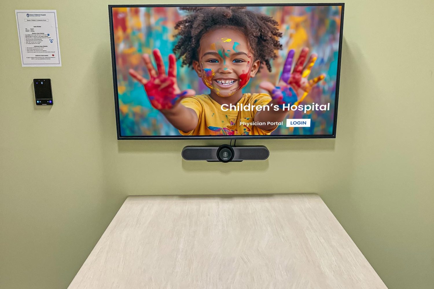 A small, enclosed meeting room with a rounded conference table, three chairs, and a wall-mounted screen displaying an image of a smiling child with painted hands. A video conferencing camera is installed below the screen.