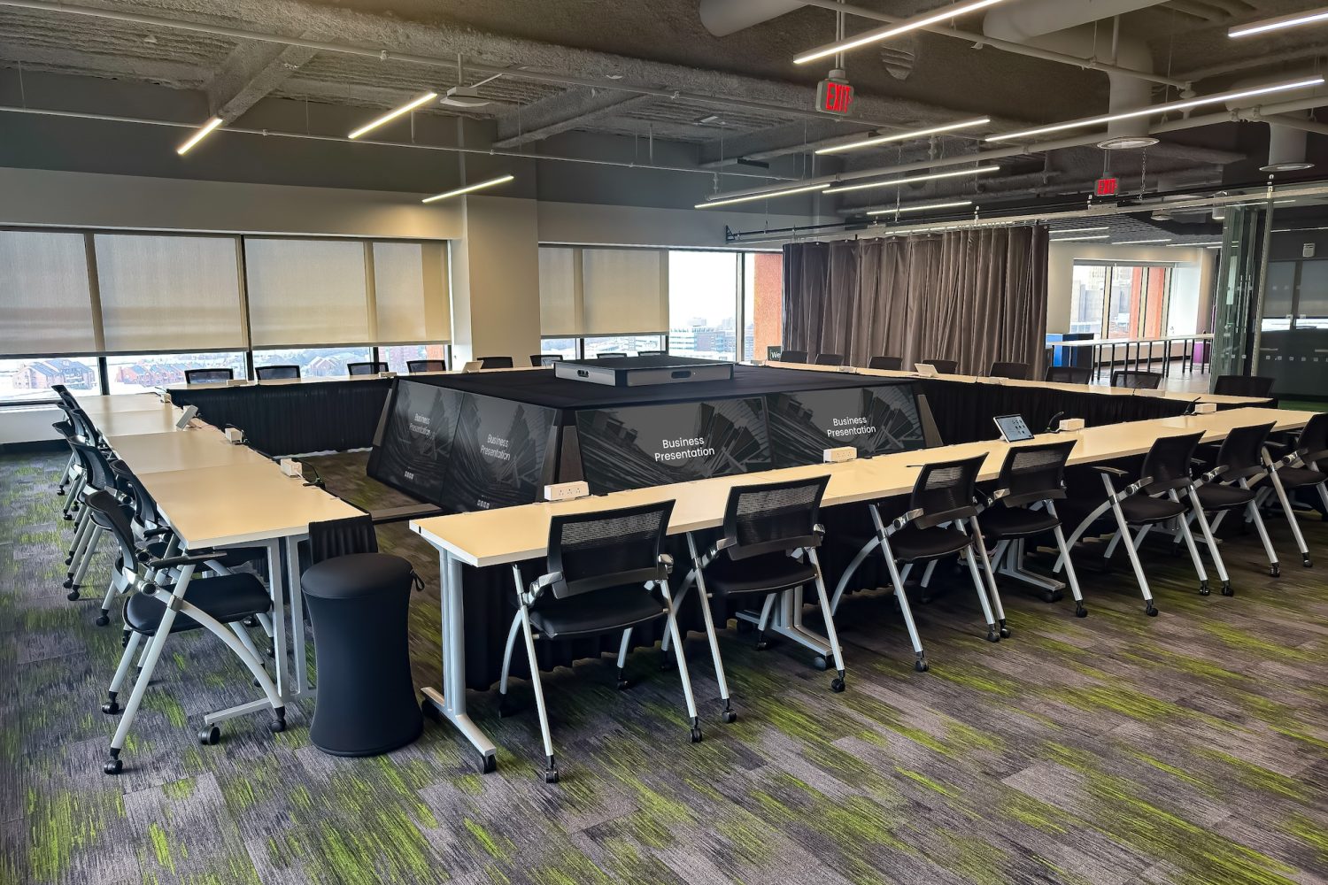 A modern executive briefing center set up in a U-shaped conference table arrangement. Large monitors are embedded in the center for presentations, and sleek office chairs surround the tables. Floor-to-ceiling windows provide natural lighting.