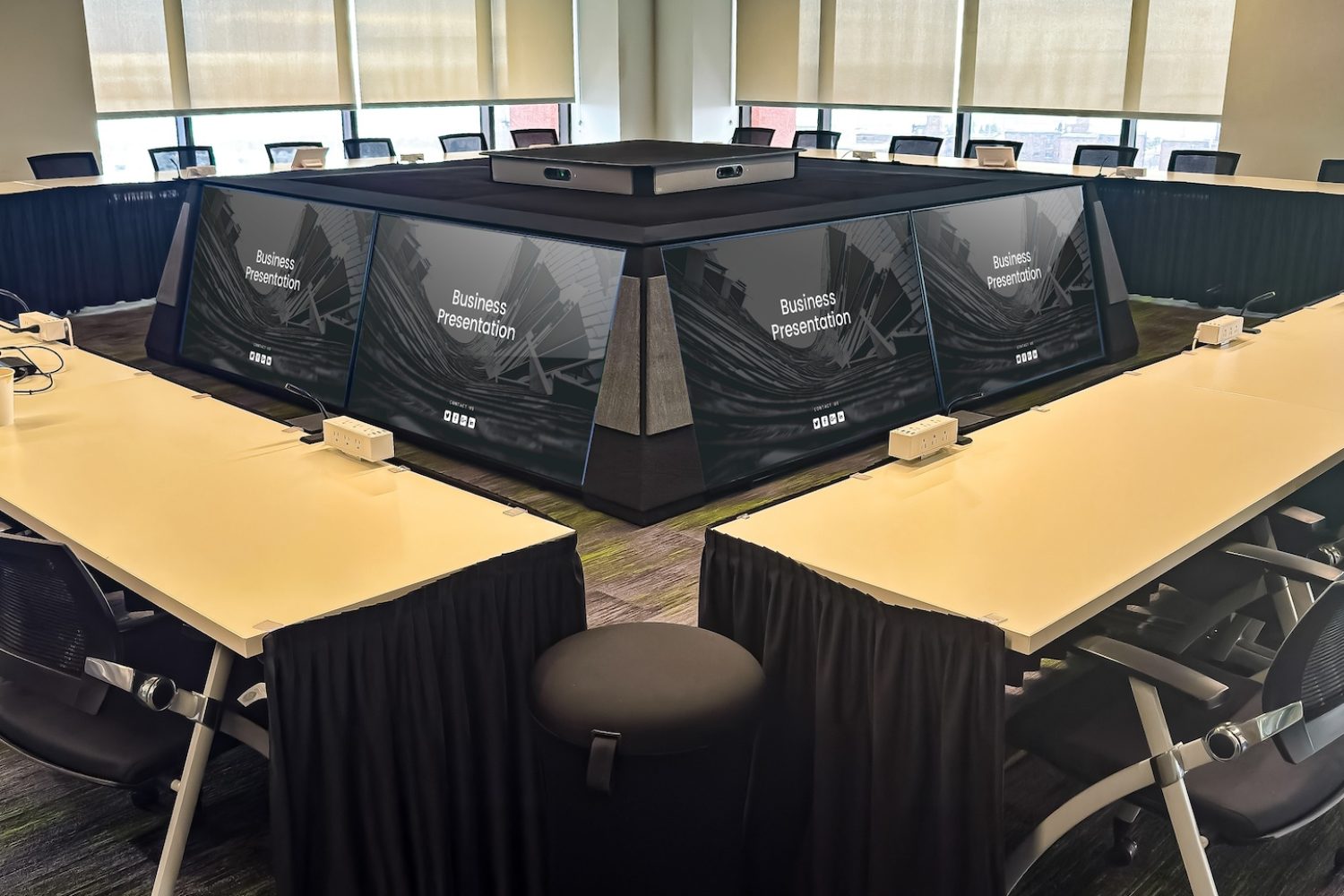 A close-up view of the central display system in the executive briefing center. Multiple screens showcase a “Business Presentation” graphic, creating an immersive meeting environment. The surrounding tables are neatly arranged with power outlets and nameplates.