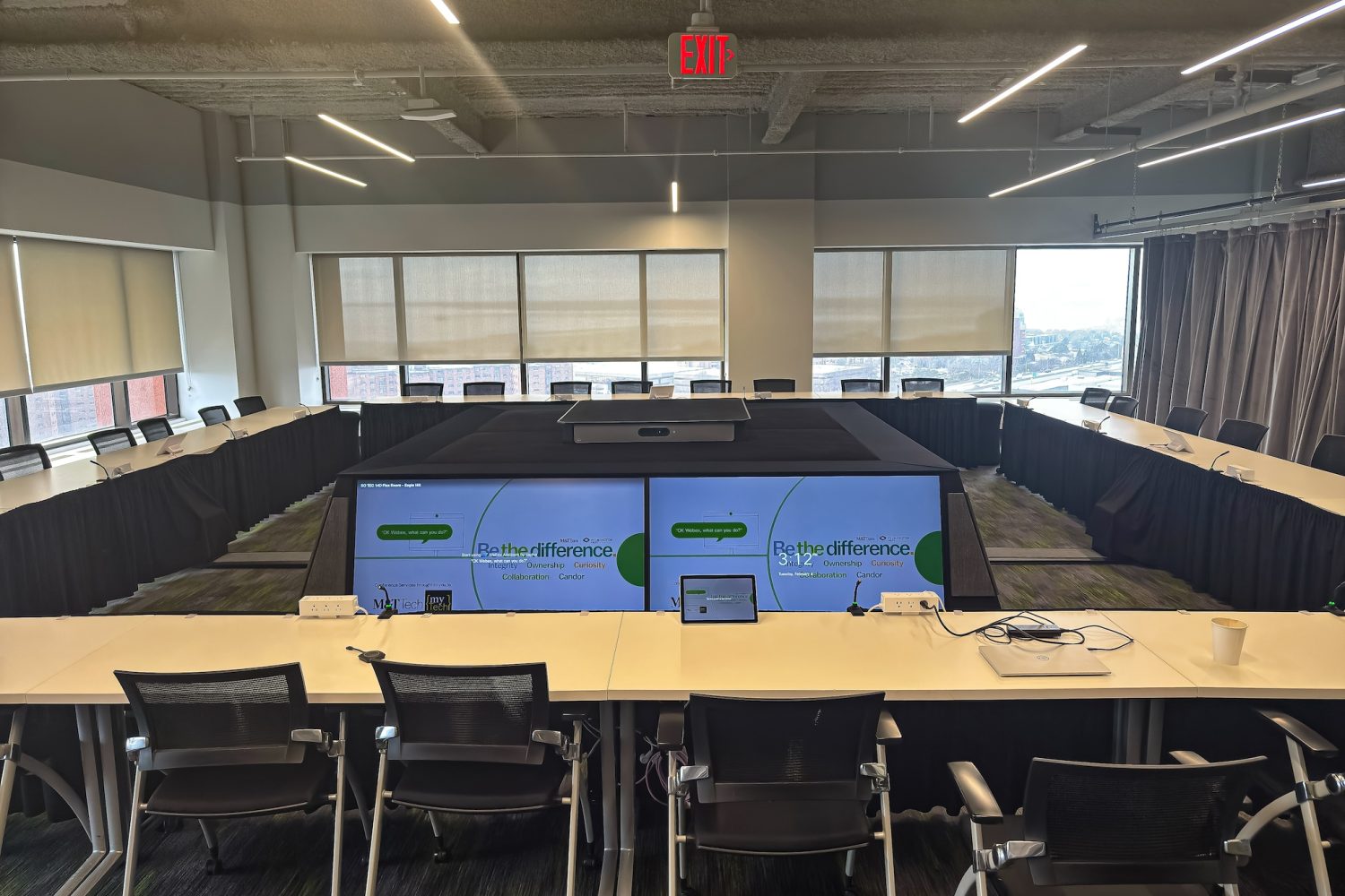 The executive briefing center in use, displaying corporate branding and presentation content on the central screens. A laptop and tablet are positioned on the table for interactivity, and a cup sits nearby, indicating an active meeting setting.