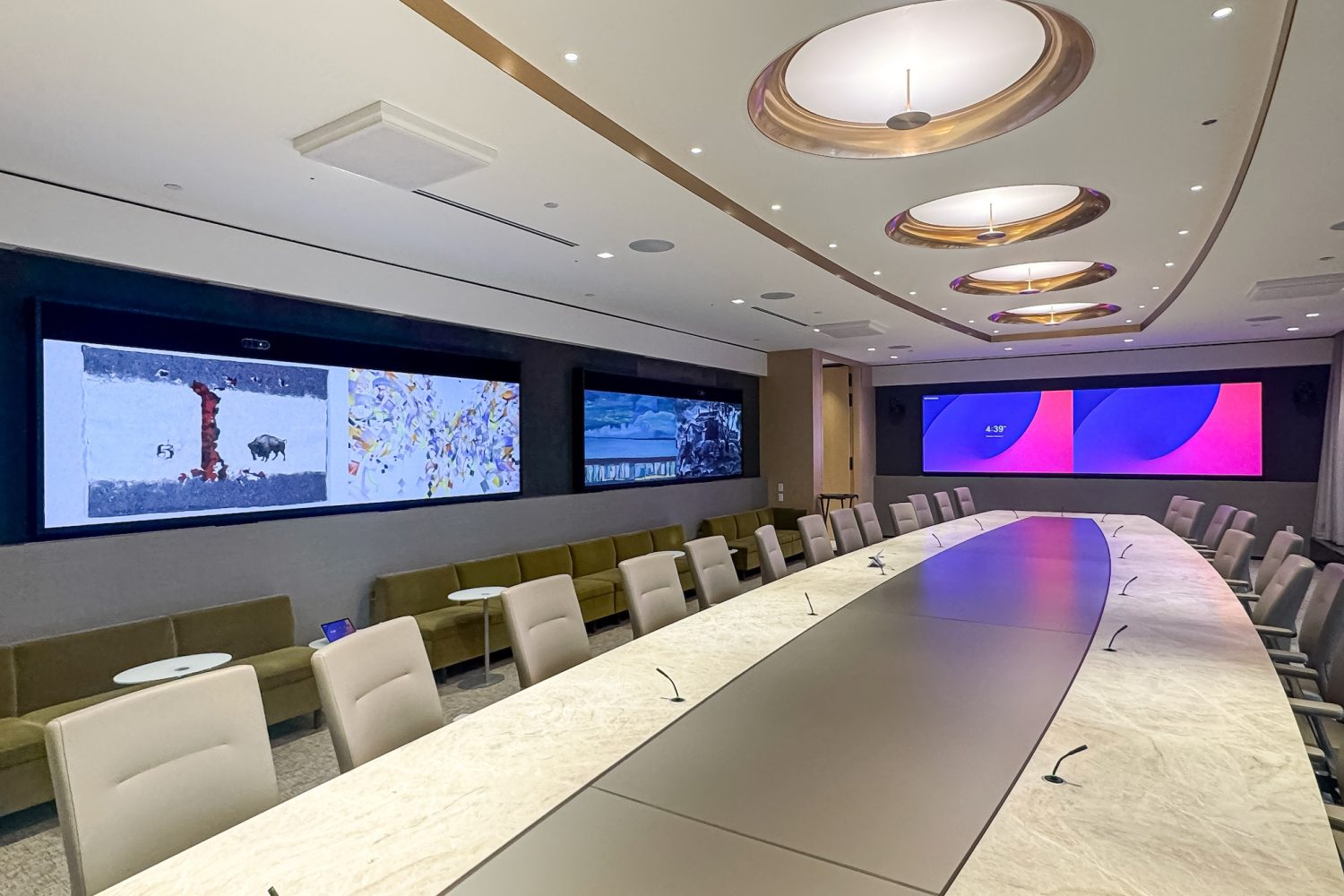 A high-tech executive boardroom featuring a long conference table with built-in microphones. The room is lined with large wall-mounted screens displaying artwork and presentations, enhancing both aesthetics and functionality.