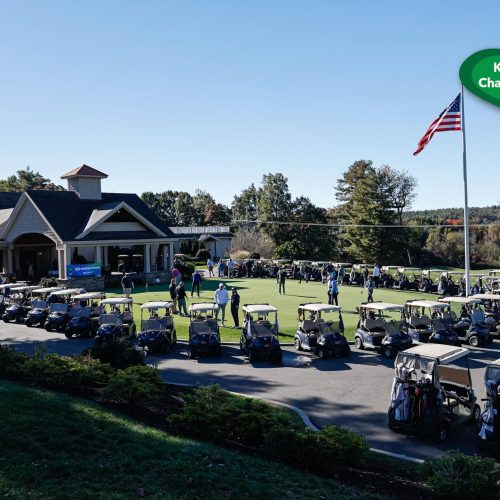 A view of a golf course with a large group of golf carts lined up outside a clubhouse, with people gathered near the entrance and an American flag flying on a tall flagpole under a clear sky.