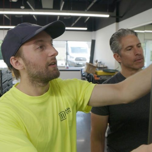 Two men in a workshop, one pointing at something on a wall.